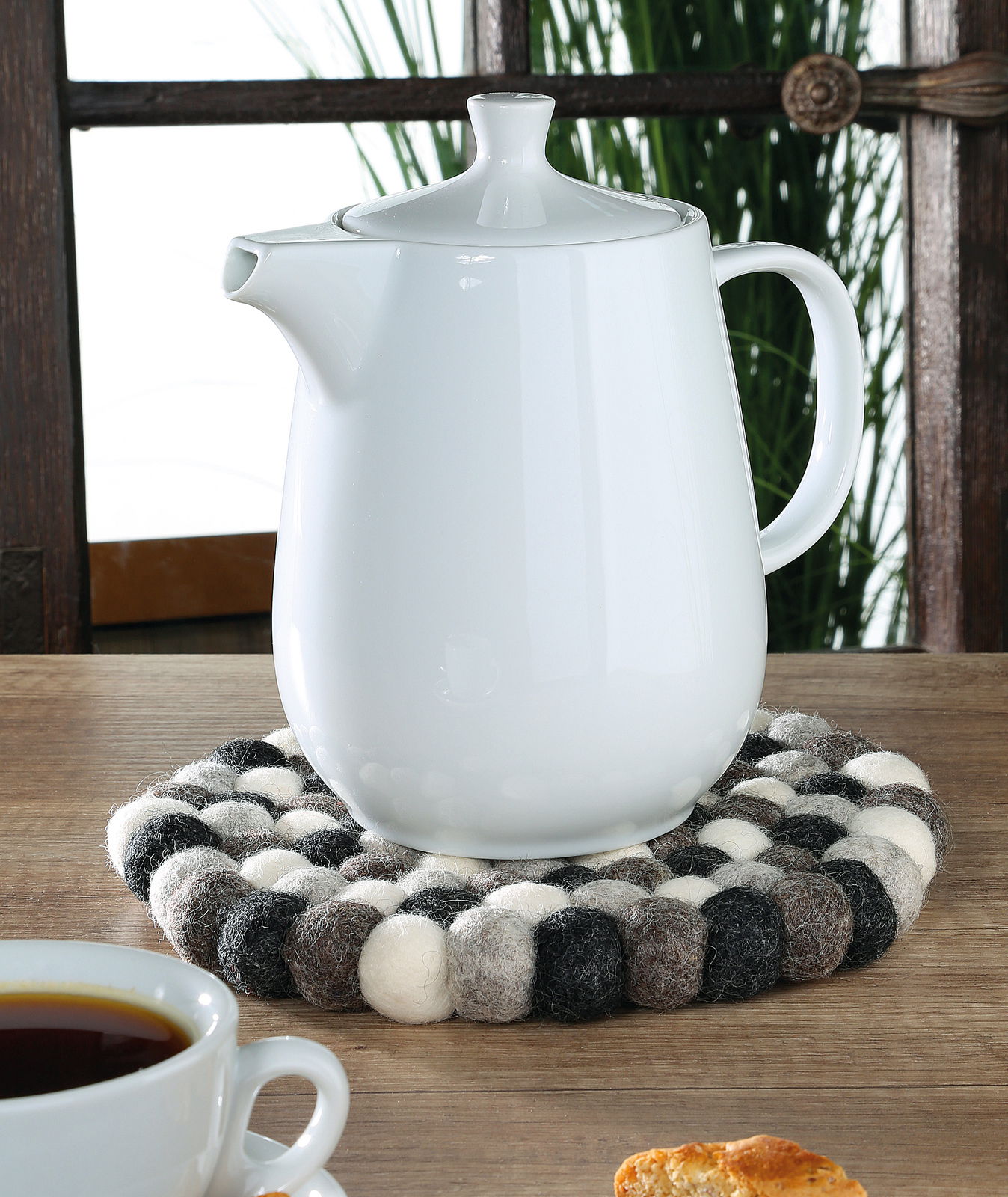 Cilio Untersetzer LANA Runder Untersetzer Lana Grigio aus Filzkugeln in Grau- und Weißtönen, auf einem Holztisch mit einer weißen Teekanne darauf, seitliche Perspektive.