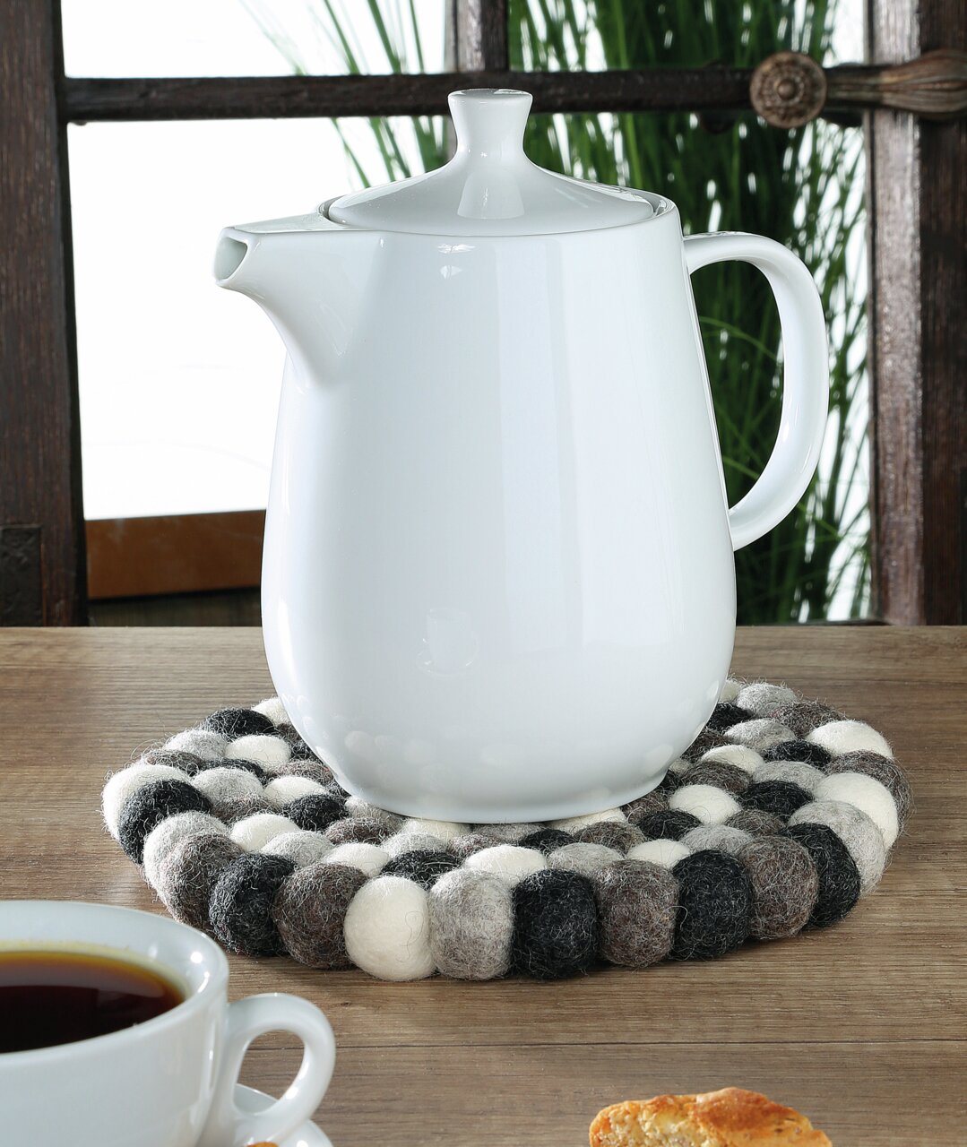 Runder Untersetzer Lana Grigio aus Filzkugeln in Grau- und Weißtönen, auf einem Holztisch mit einer weißen Teekanne darauf, seitliche Perspektive.
