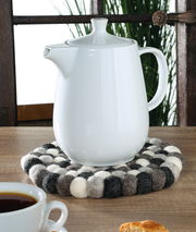 Runder Untersetzer Lana Grigio aus Filzkugeln in Grau- und Weißtönen, auf einem Holztisch mit einer weißen Teekanne darauf, seitliche Perspektive.