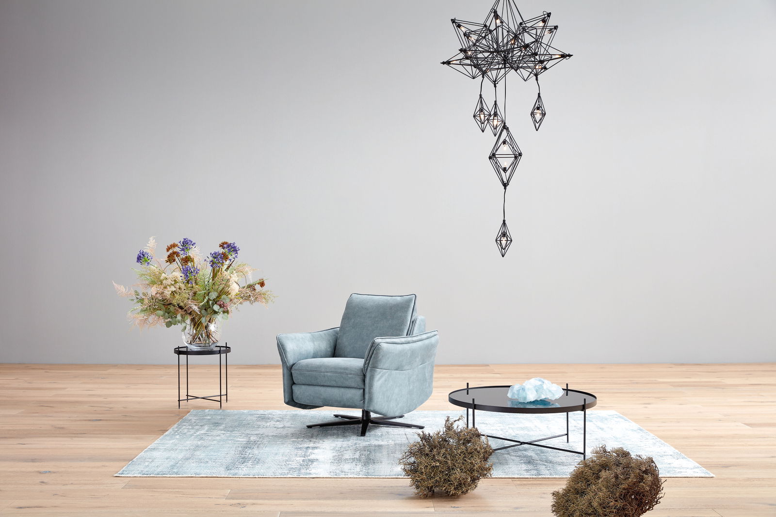 Moderner Relaxsessel in hellblauem Stoff, frontal fotografiert, auf einem hellen Holzboden mit Teppich, neben einem Beistelltisch mit Blumen und einem Couchtisch mit Dekoration.