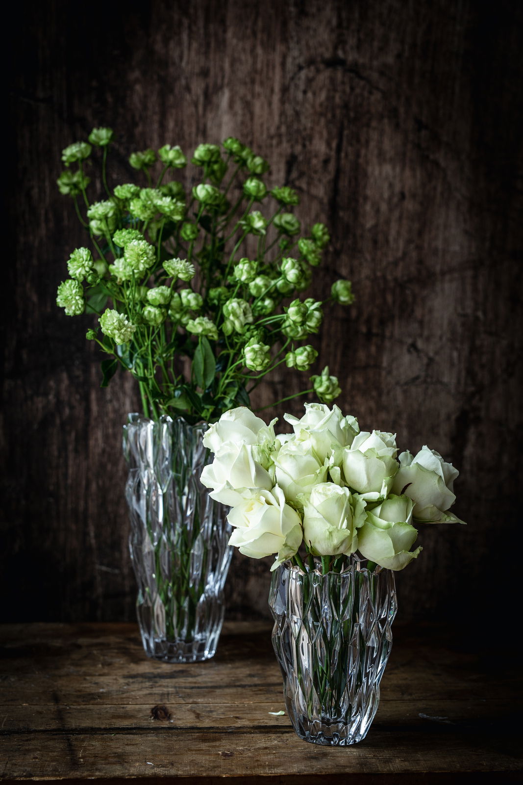 Zwei elegante Vasen aus Kristallglas, eine mit grünen Blumen und die andere mit weißen Rosen, auf einem rustikalen Holztisch vor einem dunklen Hintergrund. Die Vasen sind aus einer leicht erhöhten Perspektive fotografiert.