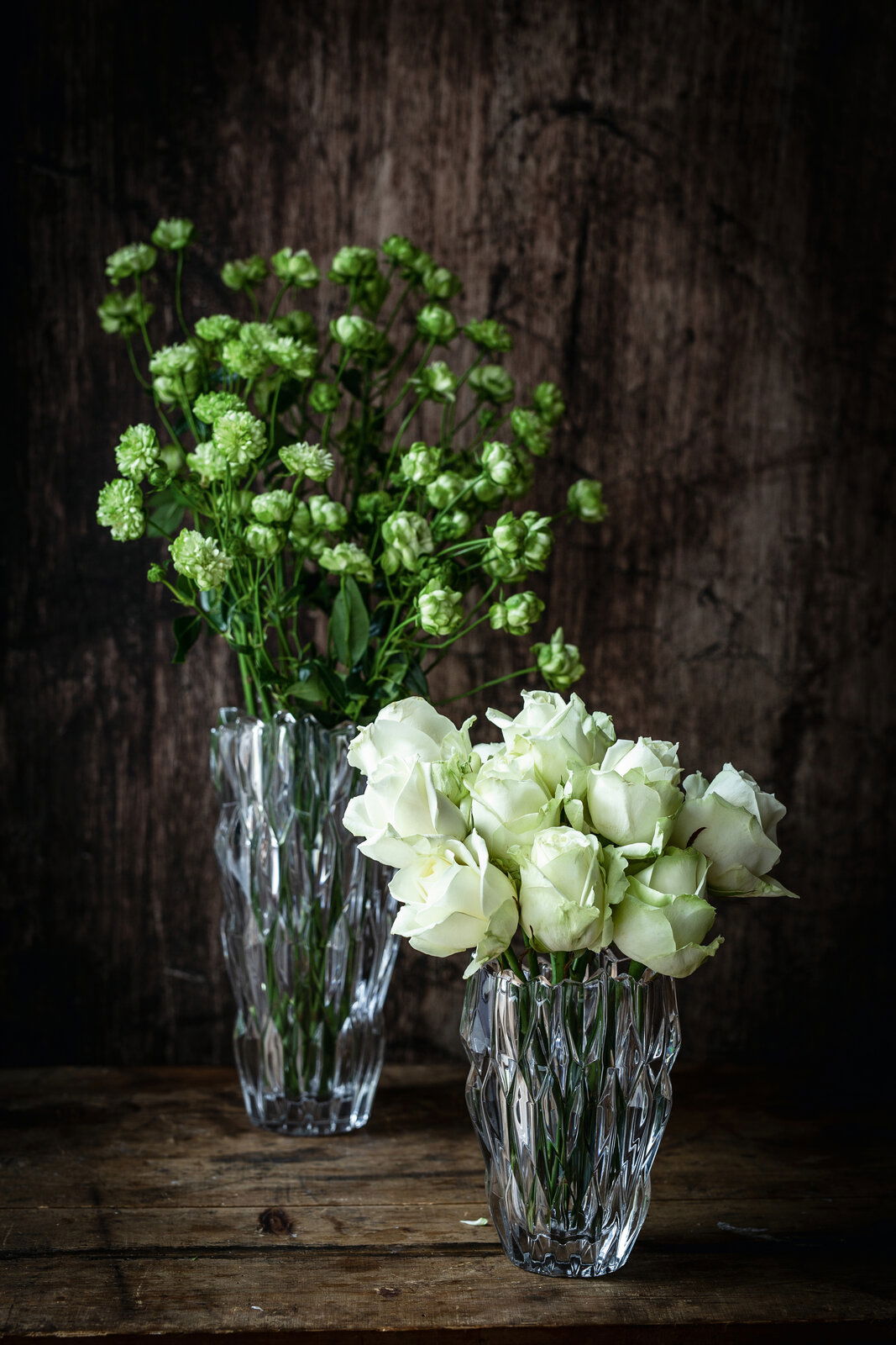 Nachtmann Vase 16cm QUARTZ Zwei elegante Vasen aus Kristallglas, eine mit grünen Blumen und die andere mit weißen Rosen, auf einem rustikalen Holztisch vor einem dunklen Hintergrund. Die Vasen sind aus einer leicht erhöhten Perspektive fotografiert.