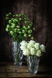 Zwei elegante Vasen aus Kristallglas, eine mit grünen Blumen und die andere mit weißen Rosen, auf einem rustikalen Holztisch vor einem dunklen Hintergrund. Die Vasen sind aus einer leicht erhöhten Perspektive fotografiert.