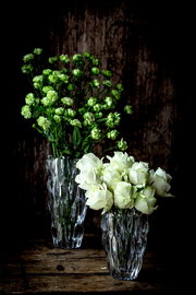 Zwei elegante Vasen aus Kristallglas, eine mit grünen Blumen und die andere mit weißen Rosen, auf einem rustikalen Holztisch vor einem dunklen Hintergrund. Die Vasen sind aus einer leicht erhöhten Perspektive fotografiert.