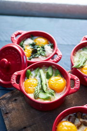 Rote Mini-Cocottes mit Deckel, gefüllt mit Eiern, Spargel und Spinat, aus der Vogelperspektive auf einem Holztisch.