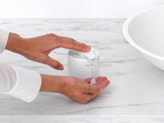Een zeepdispenser op een marmeren oppervlak, gezien van bovenaf, met een hand die zeep uit de dispenser drukt.