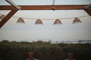 Outdoor-Lichterkette mit rustikalen Lampenschirmen aus Holz, aufgehängt an einem Holzrahmen mit Blick auf das Meer bei Sonnenuntergang.