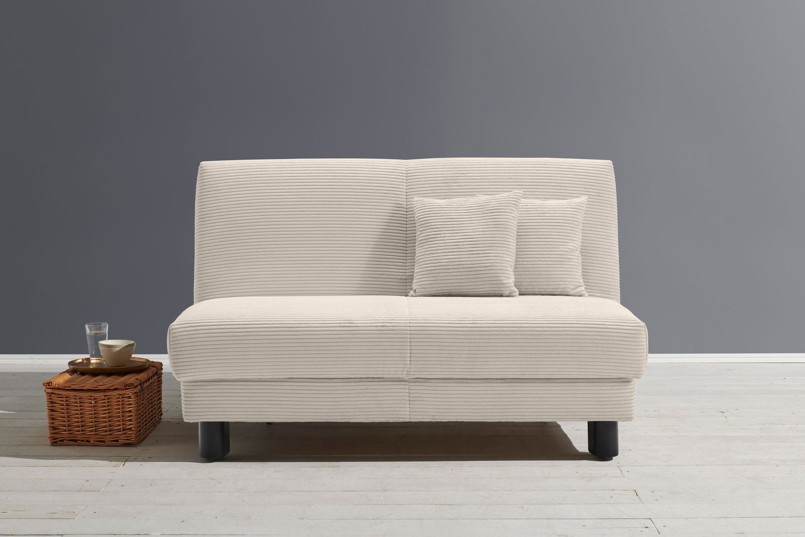 Z2 Schlafsofa ENNY Beiges Funktionssofa mit zwei passenden Kissen, frontal fotografiert, auf hellem Holzboden vor grauer Wand, neben einem Korbtisch mit Glas und Schale.