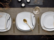 Draufsicht auf modern gedeckten Tisch mit weißen, quadratisch abgerundeten Porzellantellern mit schwarzen und goldenen Linien, goldenem Besteck, Weinglas und Kerzen auf Leinentischläufer.