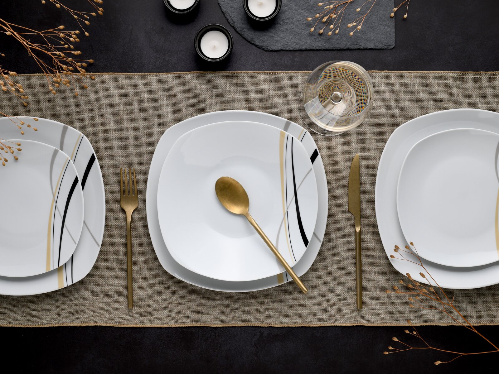 Draufsicht auf modern gedeckten Tisch mit weißen, quadratisch abgerundeten Porzellantellern mit schwarzen und goldenen Linien, goldenem Besteck, Weinglas und Kerzen auf Leinentischläufer.