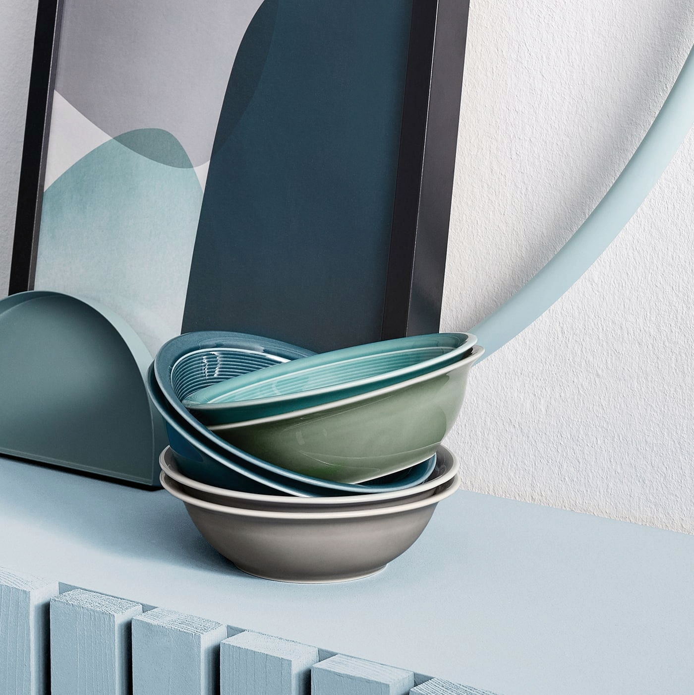 Stapel von Schalen in verschiedenen Farben, darunter Grau, Grün und Blau, auf einem Tisch vor einem abstrakten Kunstwerk. Perspektive von der Seite.