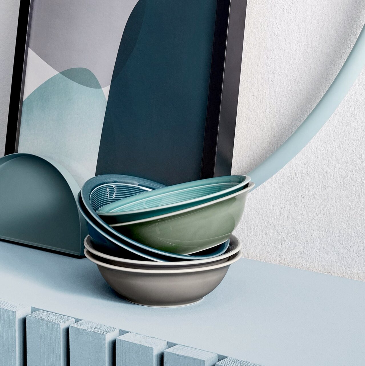 Stapel von Schalen in verschiedenen Farben, darunter Grau, Grün und Blau, auf einem Tisch vor einem abstrakten Kunstwerk. Perspektive von der Seite.