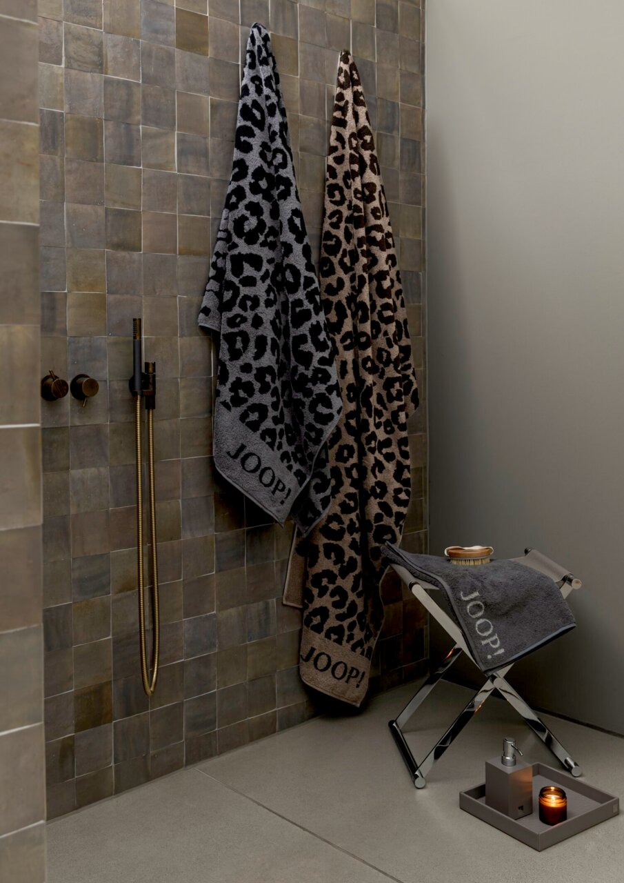 JOOP! Saunatuch LEO Zwei JOOP! Saunatücher mit Leopardenmuster, eines in Grau und eines in Braun, hängen an einer Wand in einem stilvollen Badezimmer. Ein weiteres graues Handtuch liegt auf einem Hocker. Perspektive: seitlich.