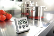 Digitaler Timer auf einer Küchenarbeitsfläche, im Hintergrund Töpfe und Tomaten, Perspektive von vorne.