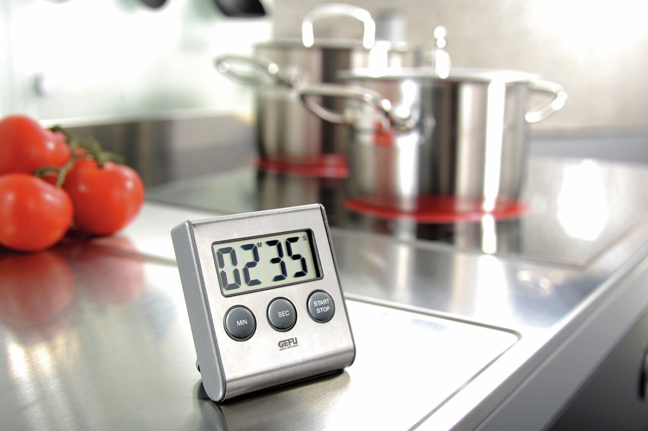 Digitaler Timer auf einer Küchenarbeitsfläche, im Hintergrund Töpfe und Tomaten, Perspektive von vorne.