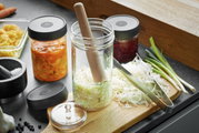 Nativo-fermentatieset op een aanrechtblad, met glazen potten, een houten stamper en verse groenten, gezien vanuit vogelperspectief.