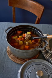 Runder Bräter Cocotte 24cm in Grau, gefüllt mit Eintopf, auf einem Tisch von oben fotografiert.