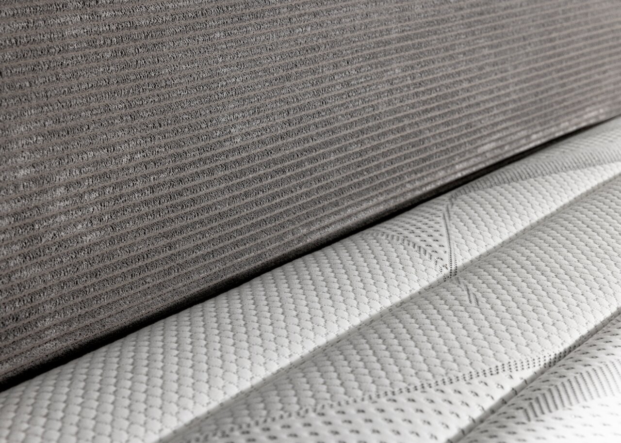 Nahaufnahme eines Boxspringbetts 180x200 mit grauem Stoffbezug und weißer, strukturierter Matratze, seitliche Perspektive