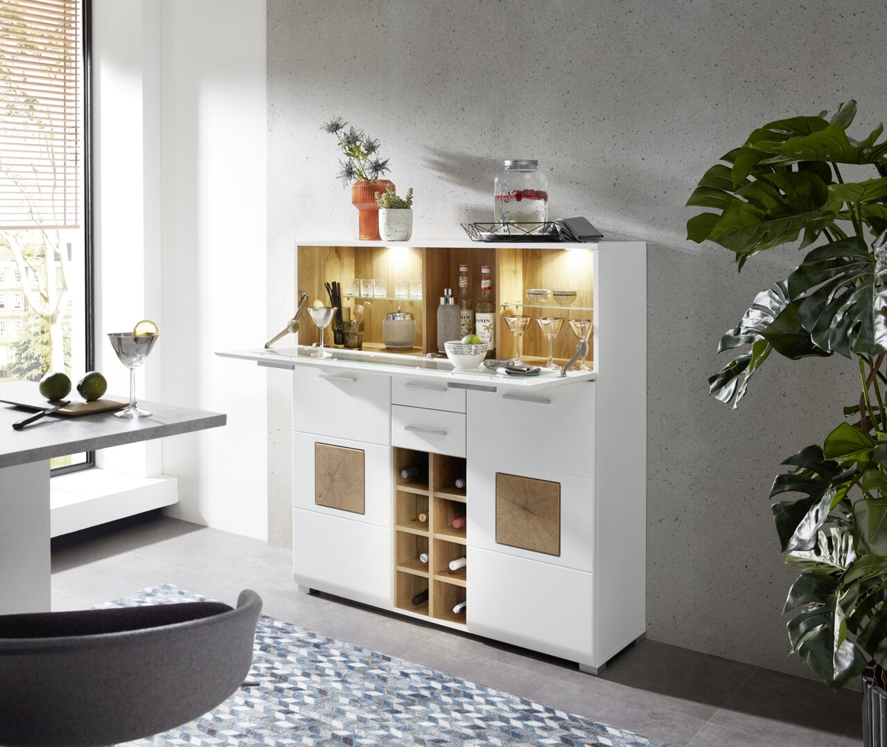 Moderner Barschrank in Weiß mit Holzdetails, gefüllt mit Gläsern und Flaschen, in einer stilvollen Wohnumgebung. Perspektive von schräg vorne.