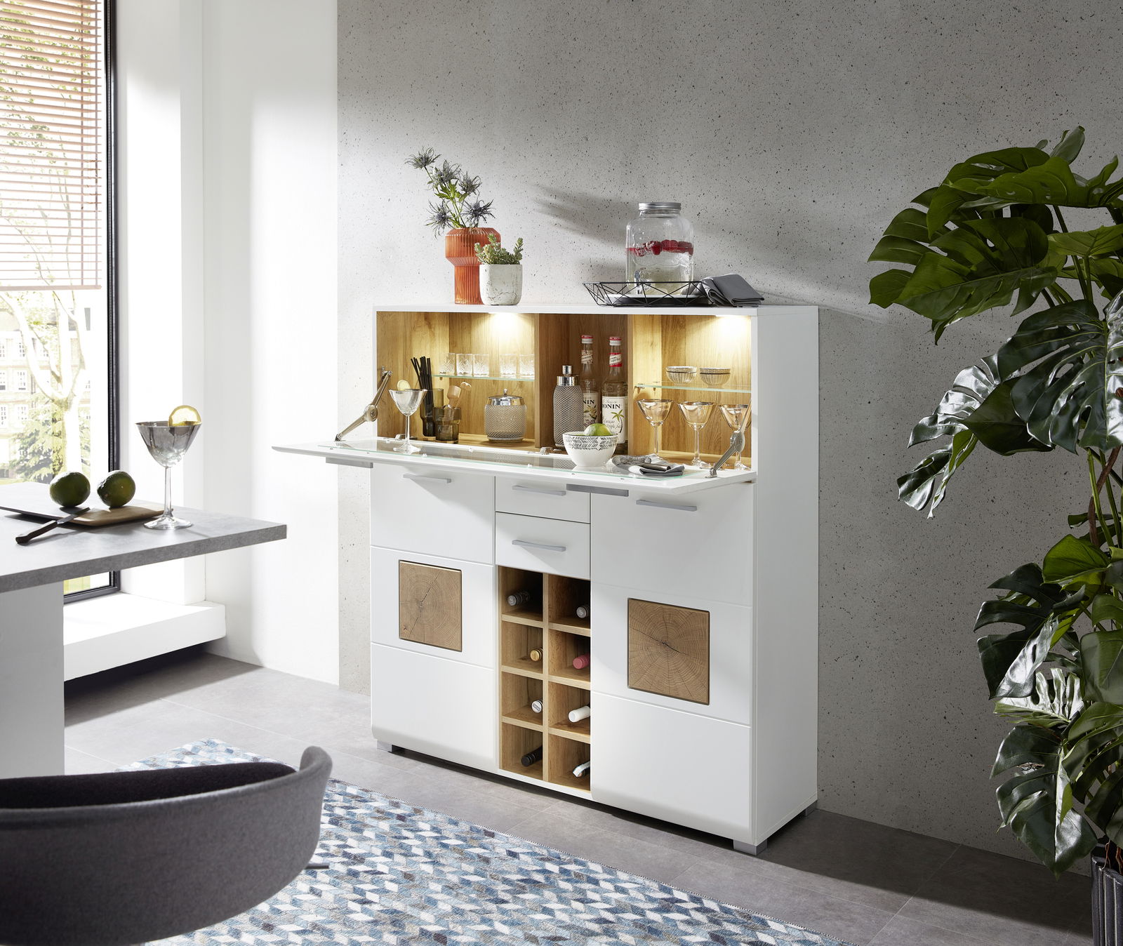 Moderner Barschrank in Weiß mit Holzdetails, gefüllt mit Gläsern und Flaschen, in einer stilvollen Wohnumgebung. Perspektive von schräg vorne.