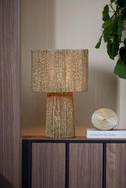 Goldene Tischuhr BOJANO mit einem Durchmesser von 15,5 cm, frontal auf einem Regal neben einer dekorativen Lampe und einem Buch platziert.