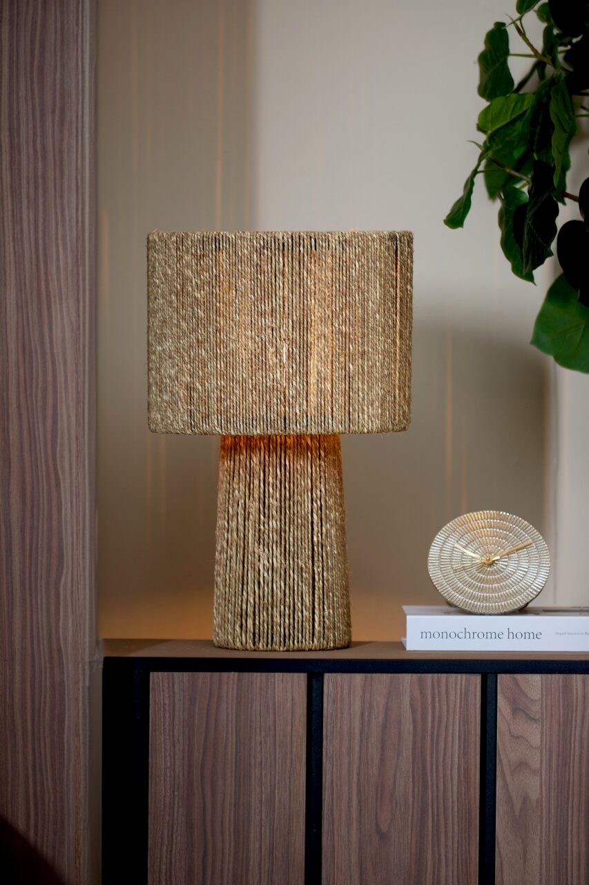 Goldene Tischuhr BOJANO mit einem Durchmesser von 15,5 cm, frontal auf einem Regal neben einer dekorativen Lampe und einem Buch platziert.