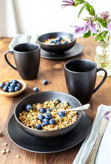 Schwarzes Geschirr-Set mit zwei Tassen und einer Schale auf einem Holztisch, gefüllt mit Müsli und Blaubeeren, aus der Vogelperspektive fotografiert.