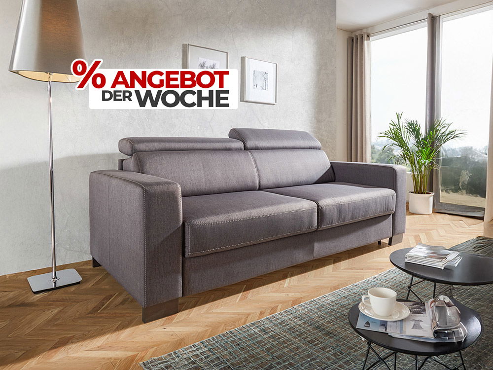 Graues 3-Sitzer Sofa mit Kopfstützen im Wohnzimmer neben Stehlampe, Couchtisch und Fensterfront