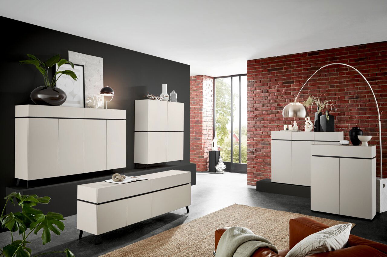 Voleo Kommode breit UNIQUE Moderne Wohnzimmermöbel in einem stilvollen Raum mit Backsteinwand, bestehend aus Kommoden und Sideboards in cremefarbenem Design, seitliche Perspektive.