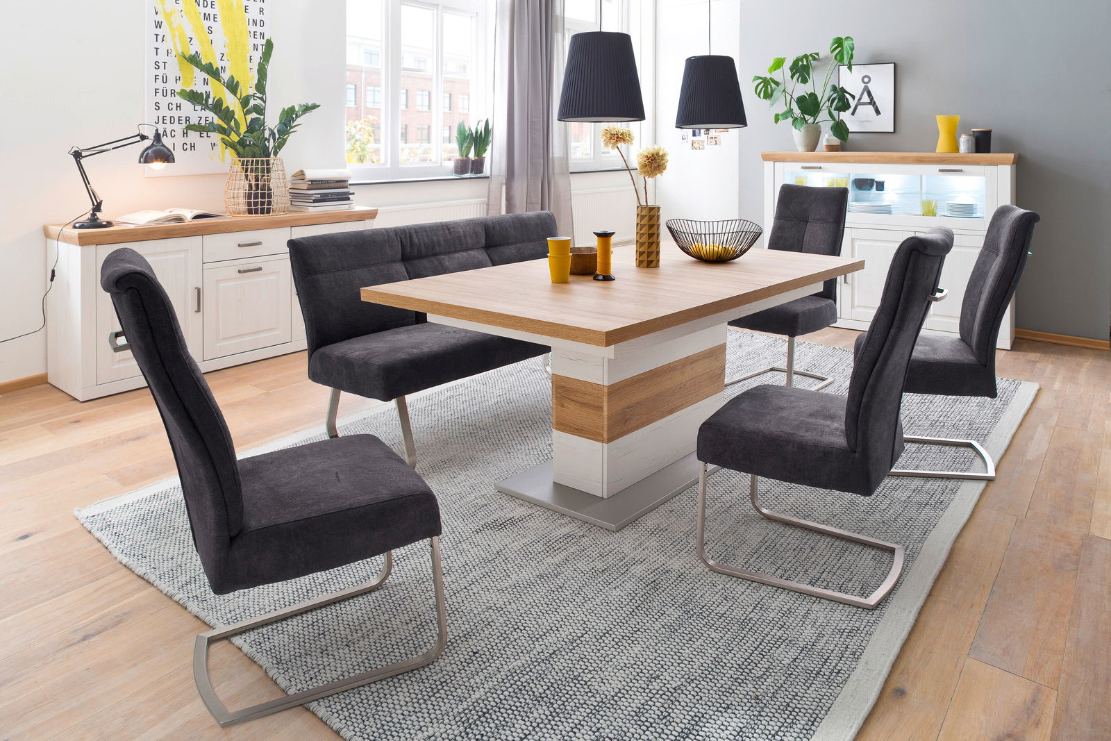 Moderner Esstisch mit Holz- und Weißakzenten, umgeben von sechs grauen Stühlen in einem hellen Esszimmer mit Pflanzen und Dekorationen, seitliche Perspektive.