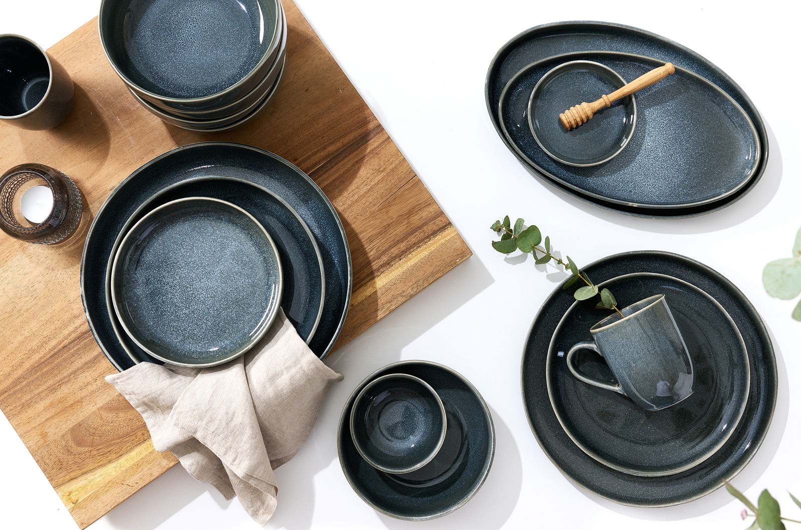 Vogelperspektive auf ein stilvolles Geschirrset in dunklem Schiefergrau, bestehend aus verschiedenen Tellern, Schalen und einer Tasse, arrangiert auf einem Holzbrett und einem weißen Untergrund, dekoriert mit einem Leinentuch und Eukalyptuszweigen.