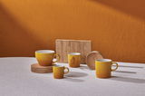 Vier gelbe Becher in verschiedenen Größen auf einem Tisch, mit einem warmen orangefarbenen Hintergrund, aus seitlicher Perspektive fotografiert.