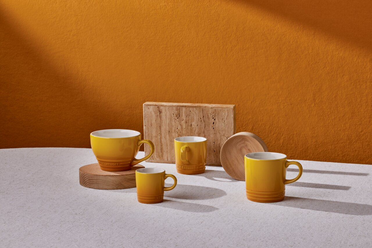 LE CREUSET Becher Nectar Vier gelbe Becher in verschiedenen Größen auf einem Tisch, mit einem warmen orangefarbenen Hintergrund, aus seitlicher Perspektive fotografiert.