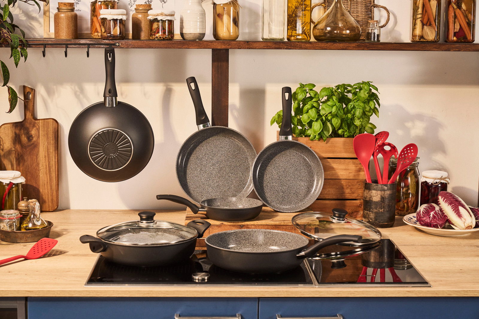 Küchenansicht mit einem Bratpfannenset, bestehend aus mehreren Pfannen mit Antihaftbeschichtung, auf einem Herd und an der Wand hängend, umgeben von Küchenutensilien und Gewürzgläsern.