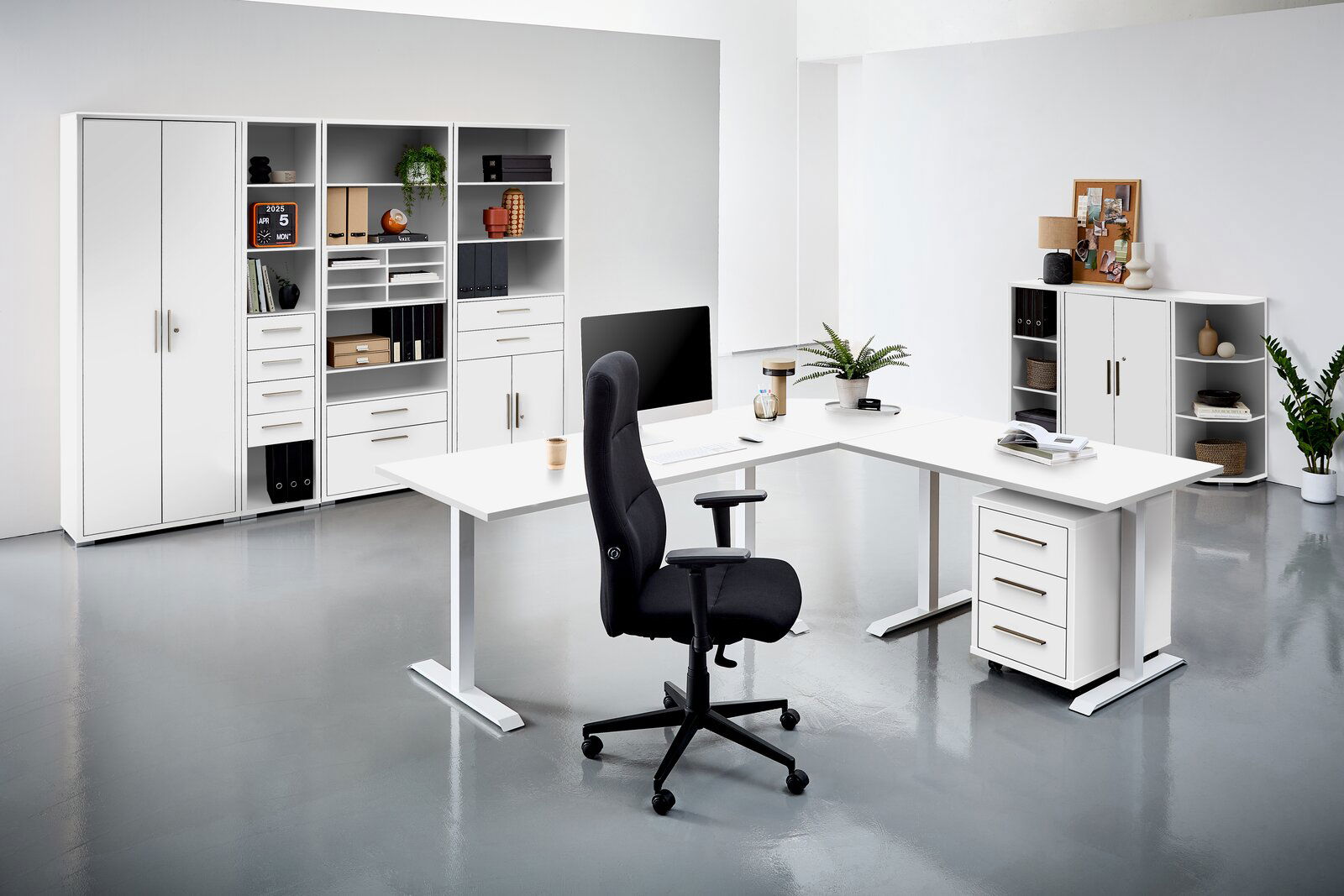 Moderne Büroumgebung mit weißen Schreibtischen, Regalen und einem schwarzen Bürostuhl, aus der Perspektive von vorne betrachtet.