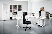 Moderne Büroumgebung mit weißen Schreibtischen, Regalen und einem schwarzen Bürostuhl, aus der Perspektive von vorne betrachtet.