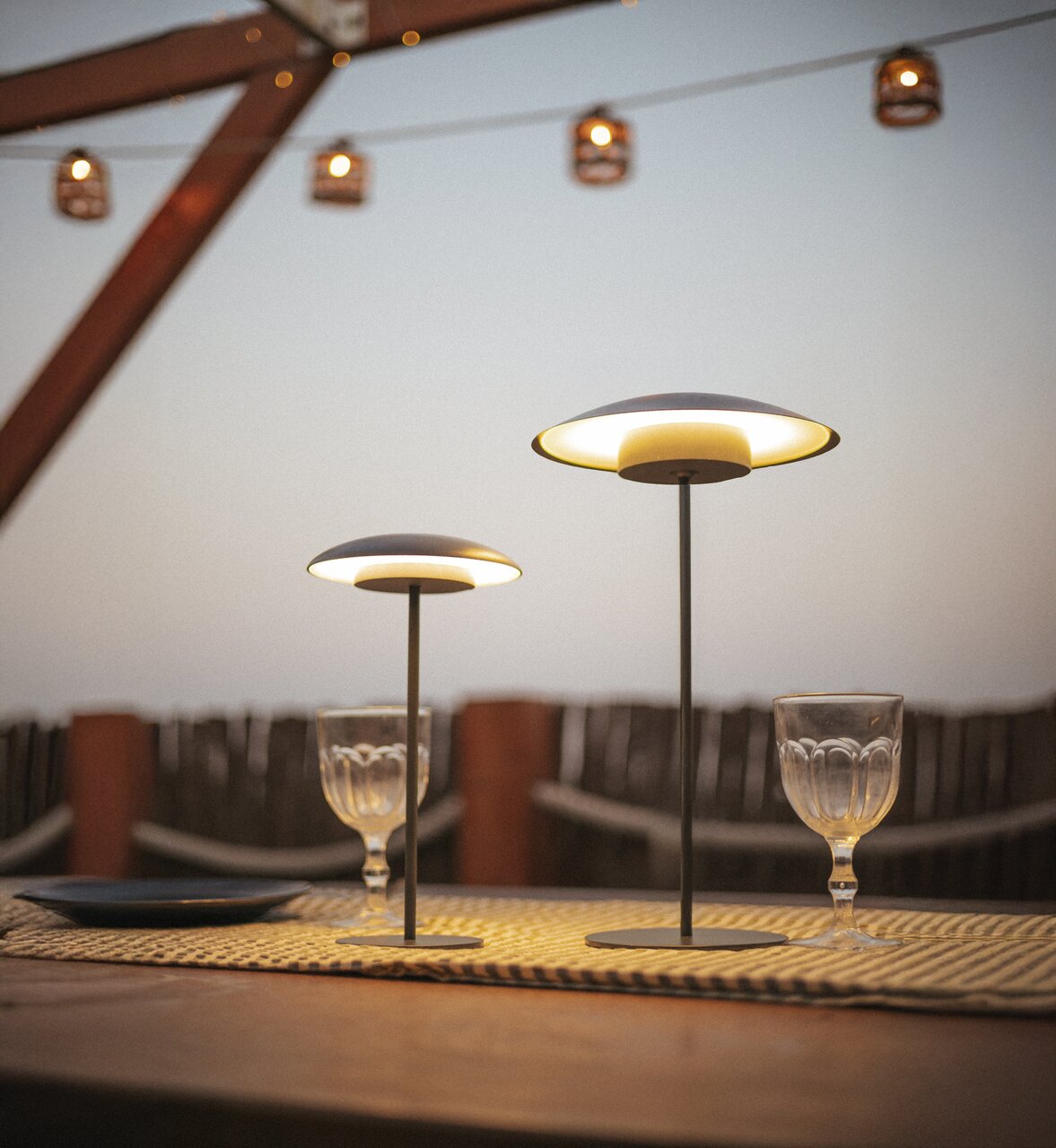 Twee moderne, draadloze led-tafellampen in verschillende maten met warmwit licht op een gedekte buitentafel, gefotografeerd vanuit een iets lager perspectief op ooghoogte bij schemering.