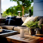 Beige, zylindrische LED-Tischleuchte mit flachem, rundem Lampenkopf auf Holztisch neben Glas-Pflanzgefäß, Outdoor-Lounge im Hintergrund; Perspektive: leicht von vorne und etwas von oben, Nahaufnahme