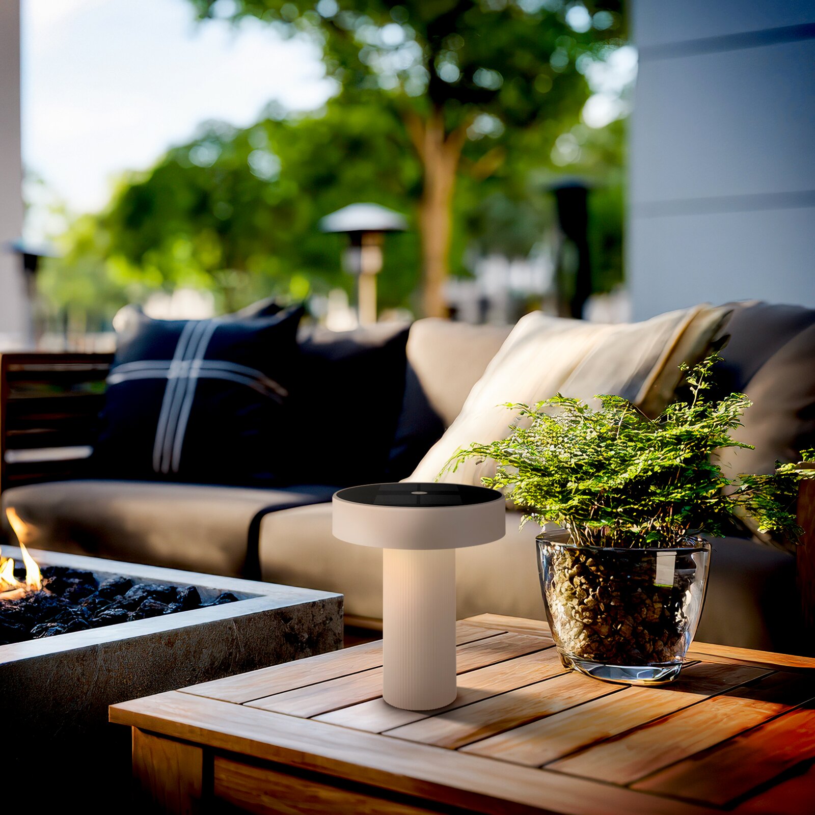 Beige, zylindrische LED-Tischleuchte mit flachem, rundem Lampenkopf auf Holztisch neben Glas-Pflanzgefäß, Outdoor-Lounge im Hintergrund; Perspektive: leicht von vorne und etwas von oben, Nahaufnahme