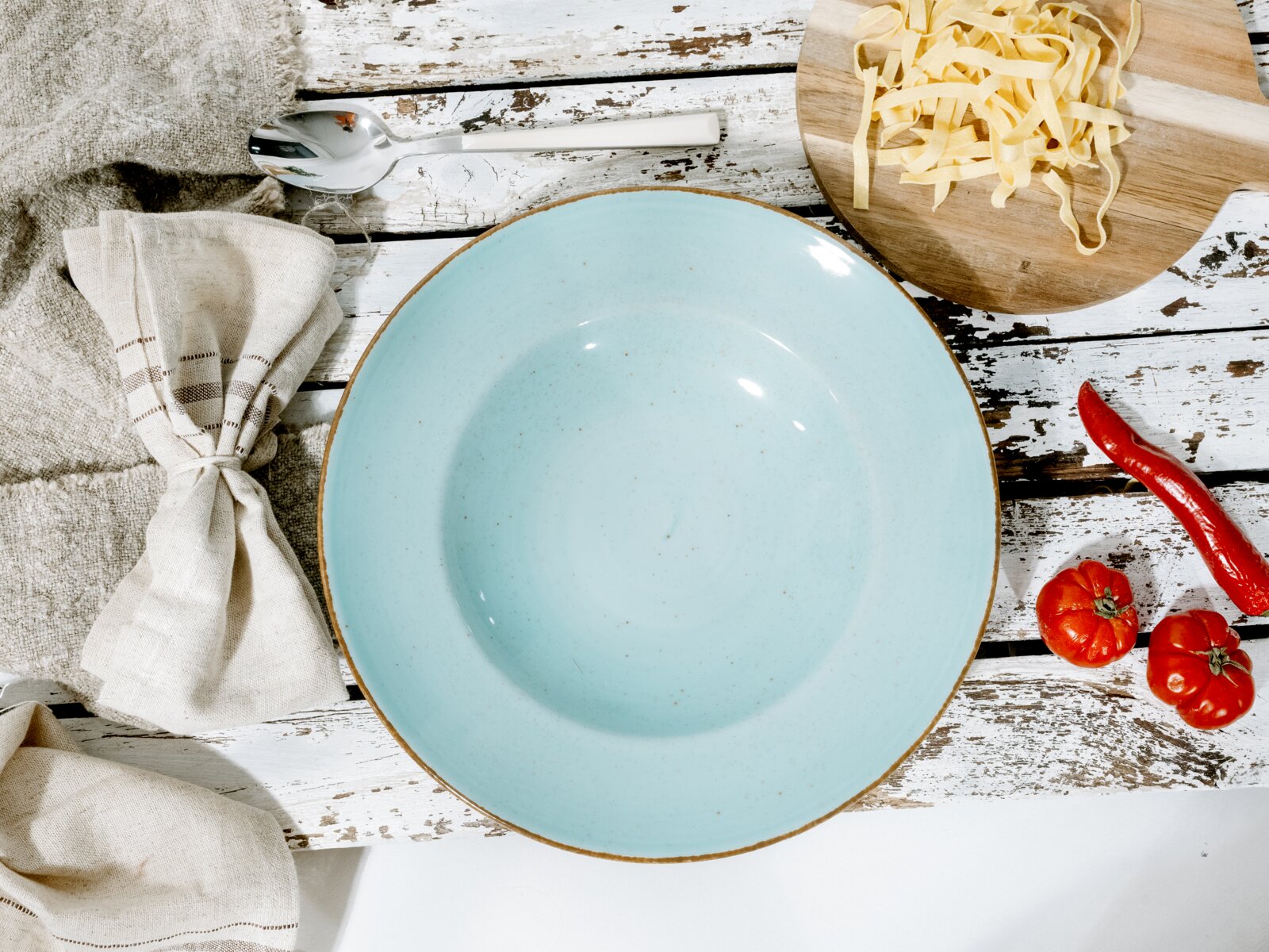 Hellblauer Keramik-Suppenteller mit braunem Rand auf rustikalem, weißem Holztisch, fotografiert in Draufsicht; daneben Löffel, Stoffserviette, Holzbrett mit Bandnudeln sowie rote Chili und Tomaten.