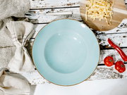 Hellblauer Keramik-Suppenteller mit braunem Rand auf rustikalem, weißem Holztisch, fotografiert in Draufsicht; daneben Löffel, Stoffserviette, Holzbrett mit Bandnudeln sowie rote Chili und Tomaten.