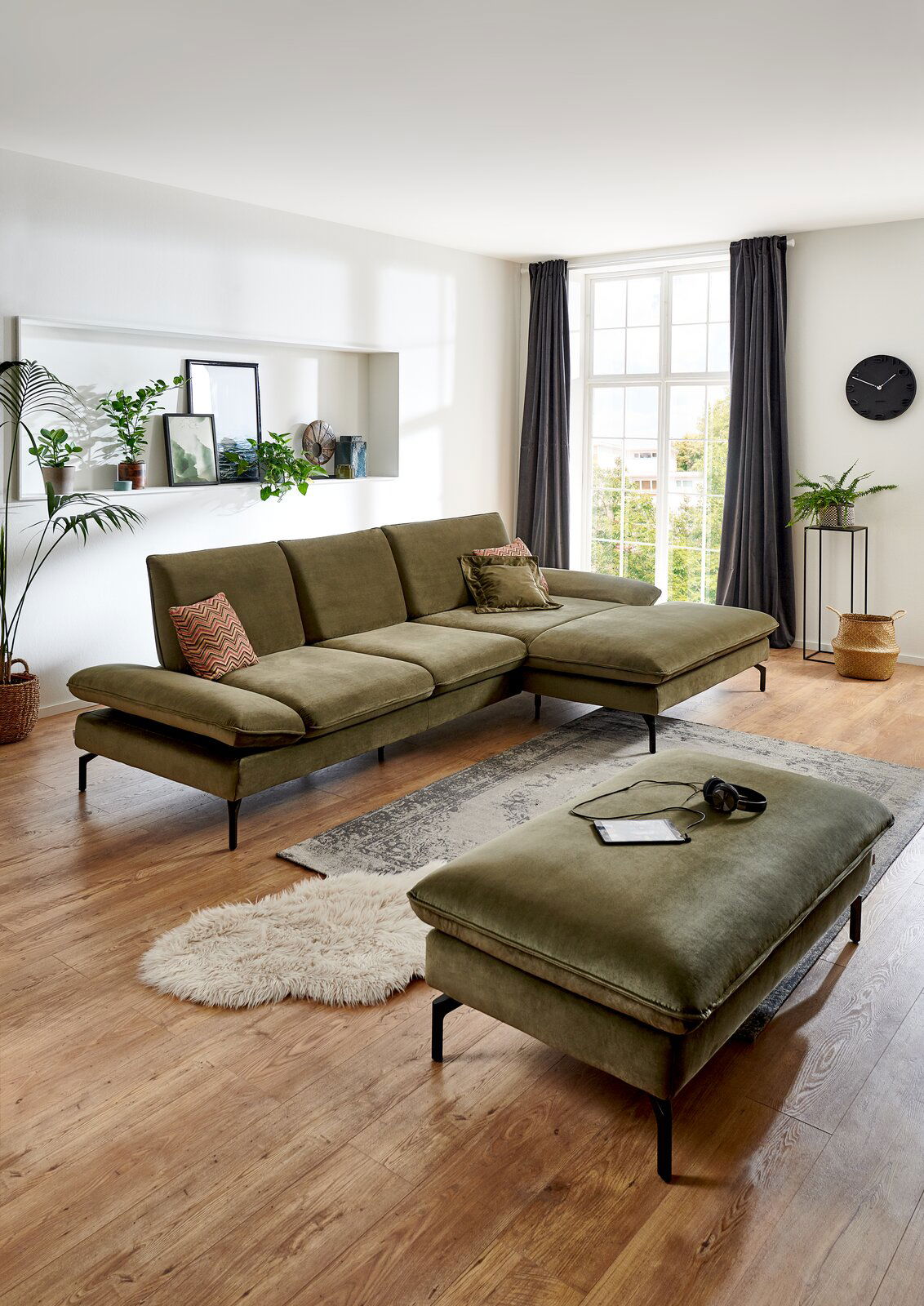 Rechteckiger Hocker in einem modernen Wohnzimmer, aus olivgrünem Stoff mit schwarzen Metallbeinen, im Vordergrund der Perspektive.