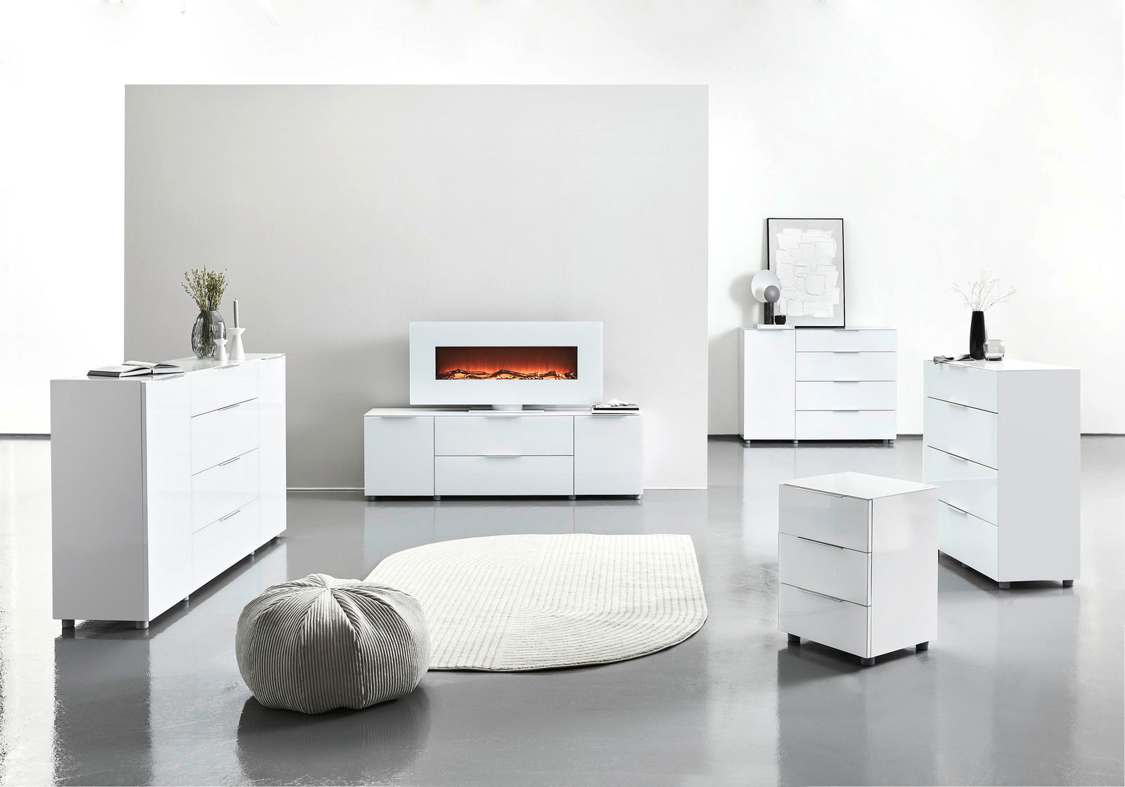 Moderne weiße Möbelkollektion in einem hellen Raum, bestehend aus Sideboards, Kommoden und einem TV-Lowboard mit elektrischem Kamin, aus der Frontalperspektive betrachtet.