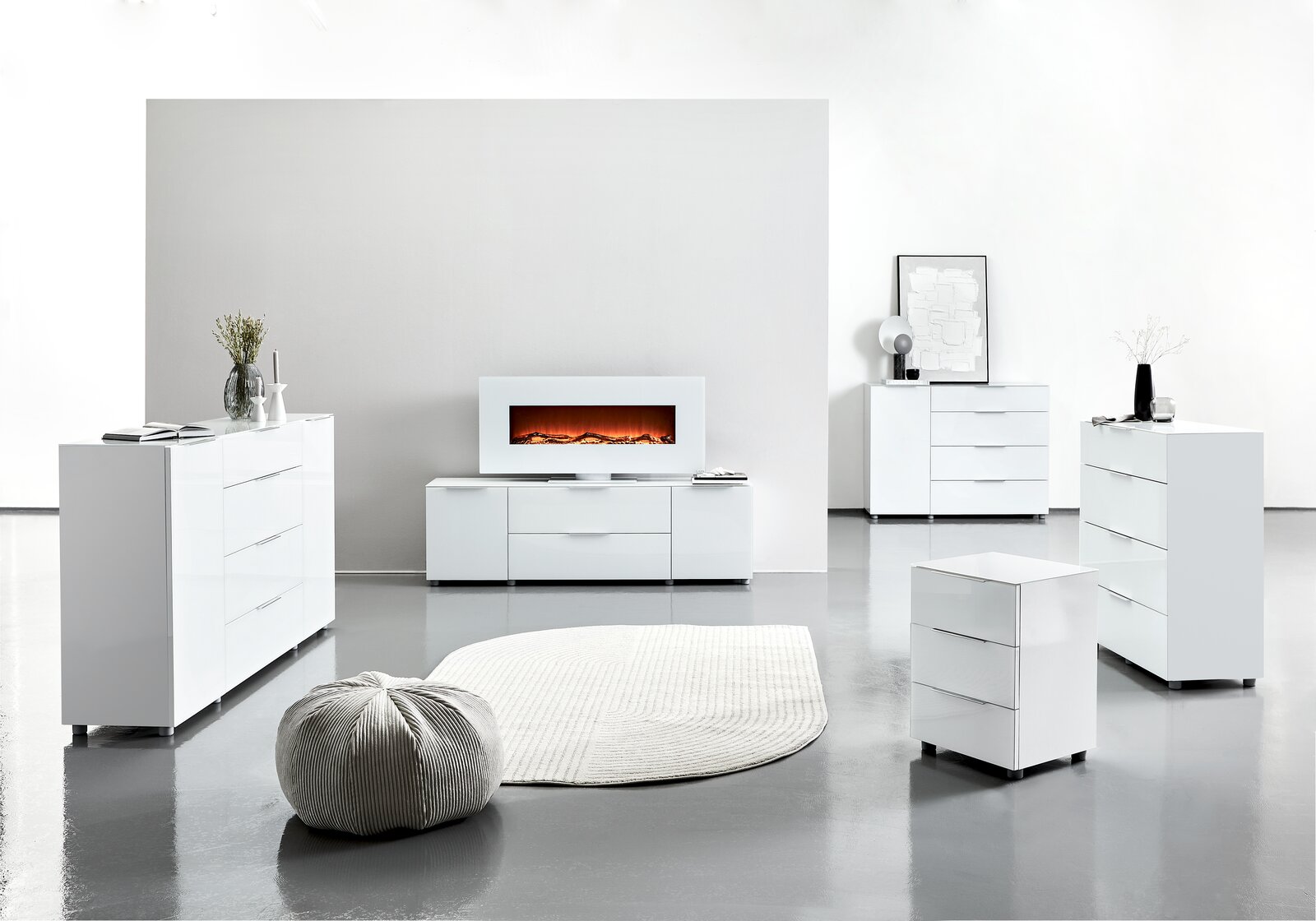 Moderne weiße Möbelkollektion in einem hellen Raum, bestehend aus Sideboards, Kommoden und einem TV-Lowboard mit elektrischem Kamin, aus der Frontalperspektive betrachtet.