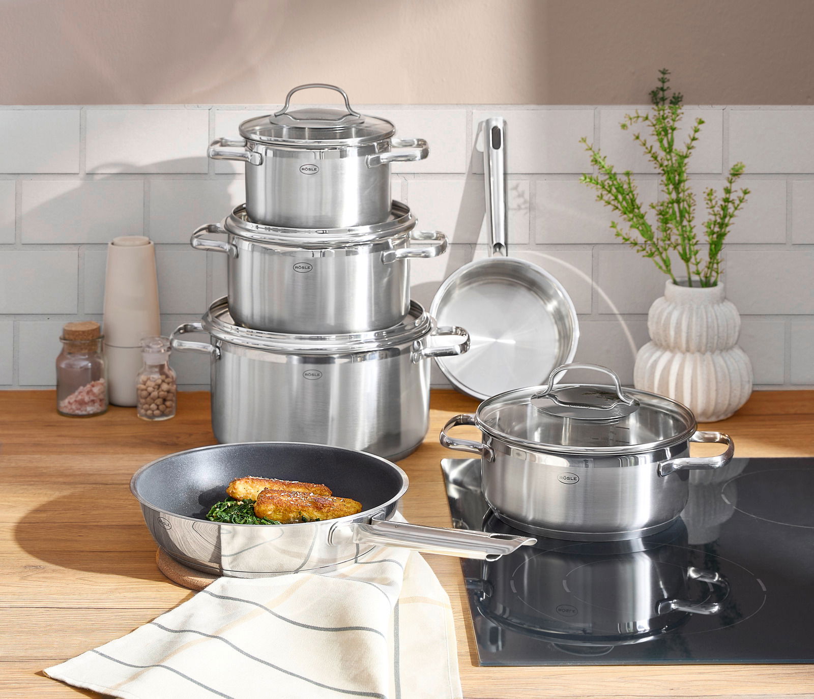 Rösle Topfset 6-tlg.ELEGANCE Topfset ELEGANCE 6-tlg. aus Edelstahl, bestehend aus mehreren Töpfen und Pfannen, auf einer Küchenarbeitsplatte präsentiert. Perspektive von vorne.