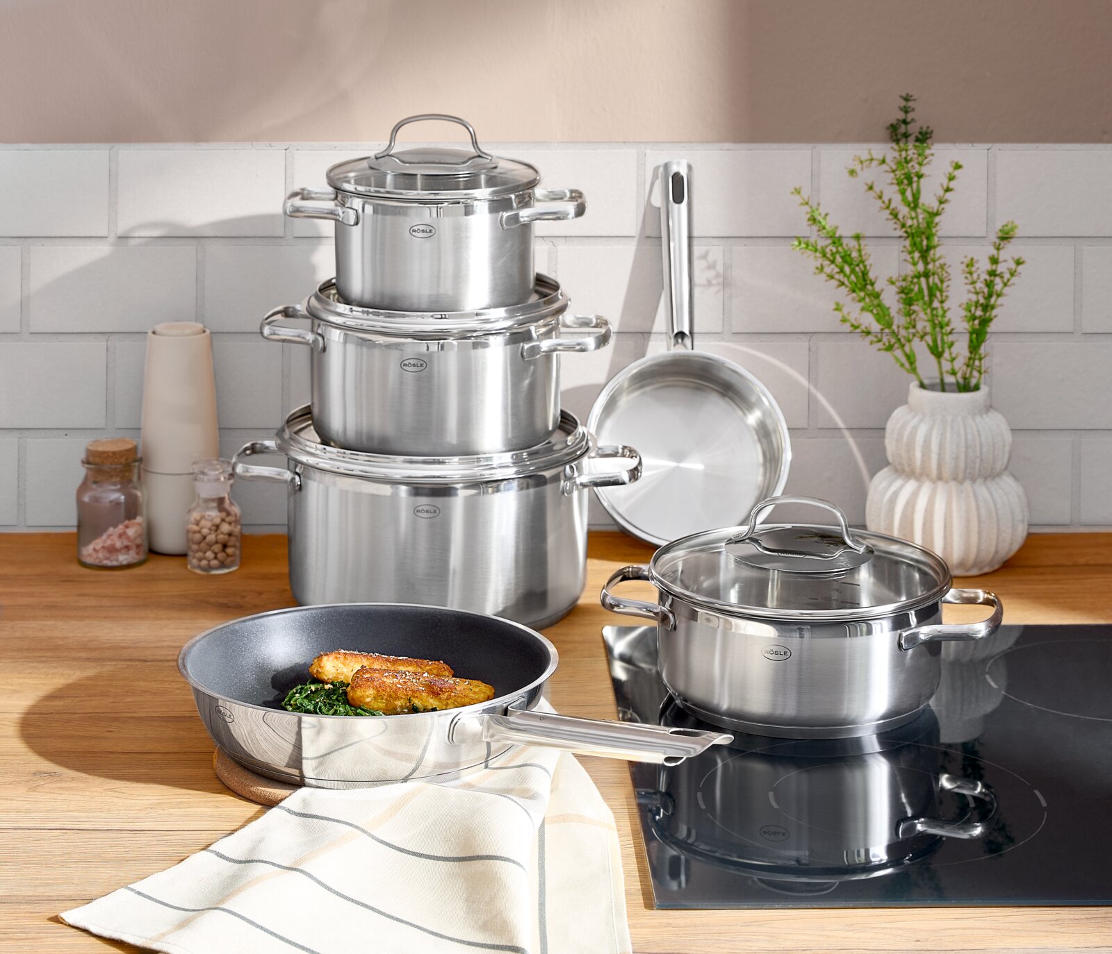 Topfset ELEGANCE 6-tlg. aus Edelstahl, bestehend aus mehreren Töpfen und Pfannen, auf einer Küchenarbeitsplatte präsentiert. Perspektive von vorne.