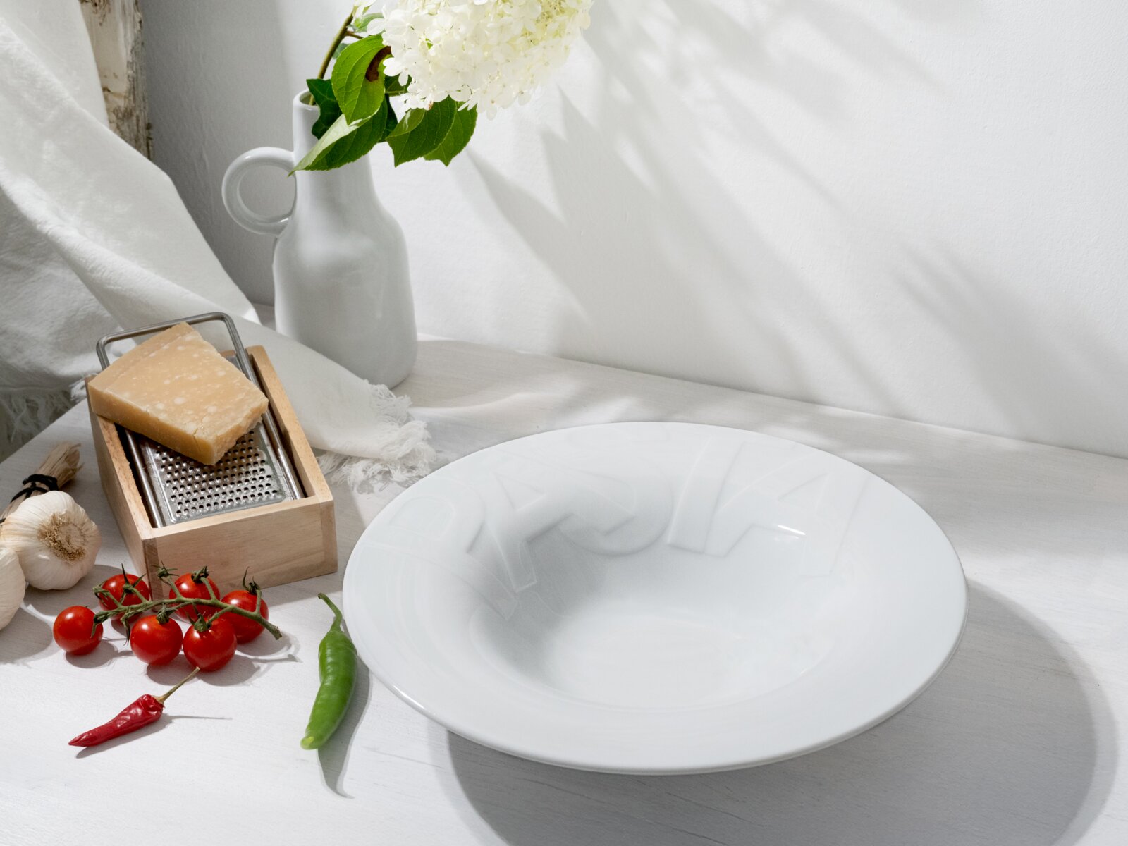Weiße, tiefe Porzellanschale auf hellem Tisch, fotografiert in leicht schräger Draufsicht; daneben Käsereibe mit Hartkäse, Kirschtomaten, Knoblauch, grüne und rote Chili sowie eine weiße Vase mit weißen Blüten im Hintergrund.