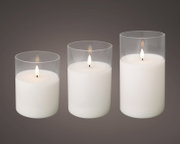 Drie witte LED-kaarsen van verschillende hoogtes in transparante glazen houders, brandend en van voren licht van bovenaf bekeken tegen een donkergrijze achtergrond