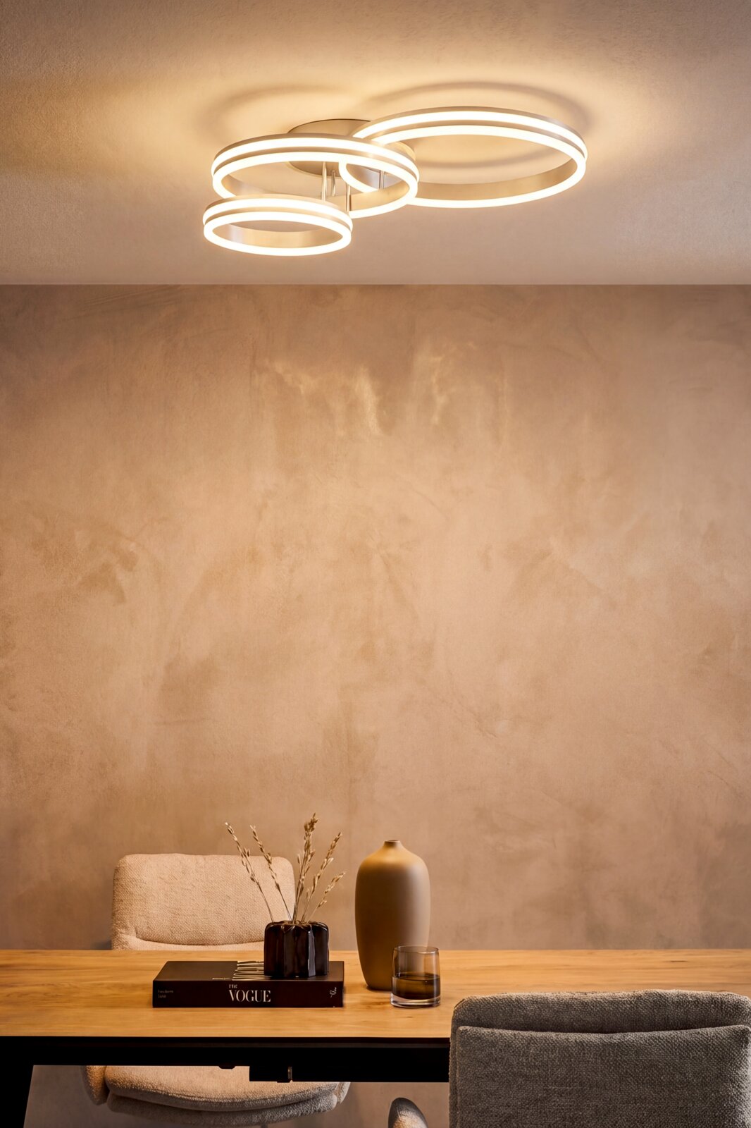 Novel LED-plafondlamp DECKENLEUCHTE Moderne led-plafondlamp met drie verlichte, elkaar overlappende ringen in wit, warmwit licht, gemonteerd aan het plafond boven een eettafel; perspectief: licht van onder naar boven (onderzicht) in de sfeer van de ruimte