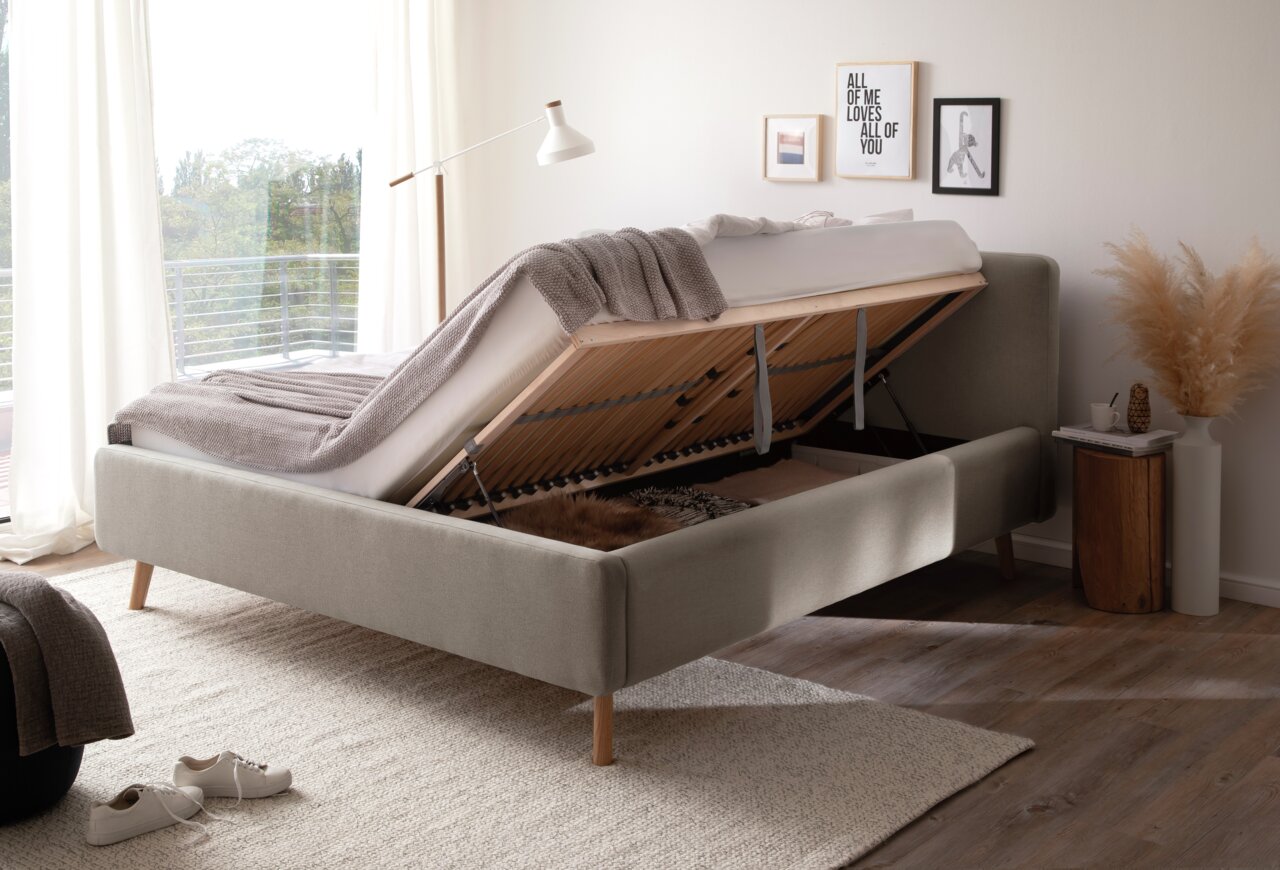 Seitliche Perspektive auf ein Polsterbett mit hochgeklappter Matratze, das Stauraum darunter zeigt. Das Bett steht in einem hellen, modern eingerichteten Raum mit Dekorationen an der Wand.
